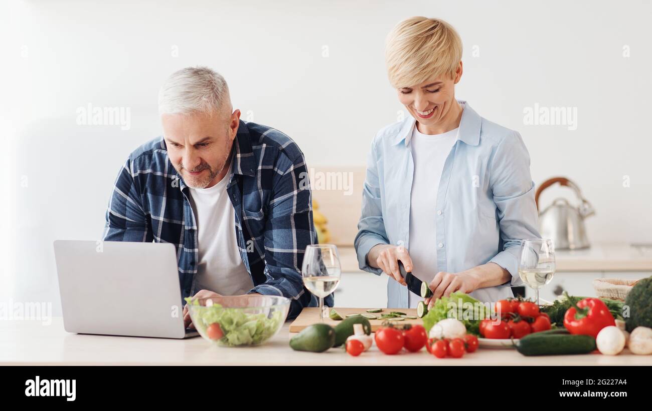 Felice coppia di mezza età 50 anni utilizzando un computer portatile, preparando cibo sano Foto Stock