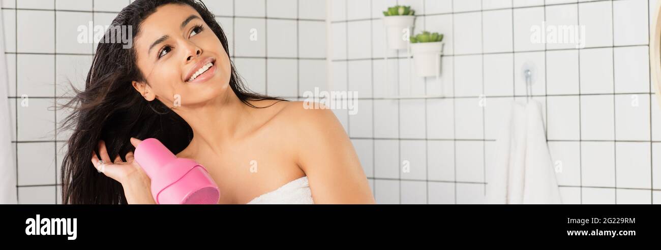 sognante giovane donna afroamericana styling capelli con asciugacapelli vicino in bagno, banner Foto Stock