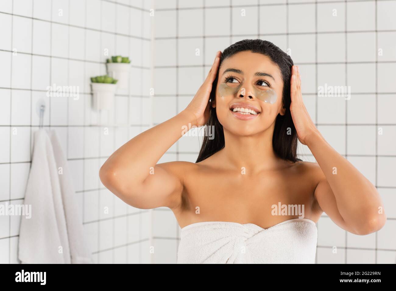 giovane donna afroamericana avvolta in un asciugamano con macchie per gli occhi che tengono le mani sulla testa in bagno Foto Stock
