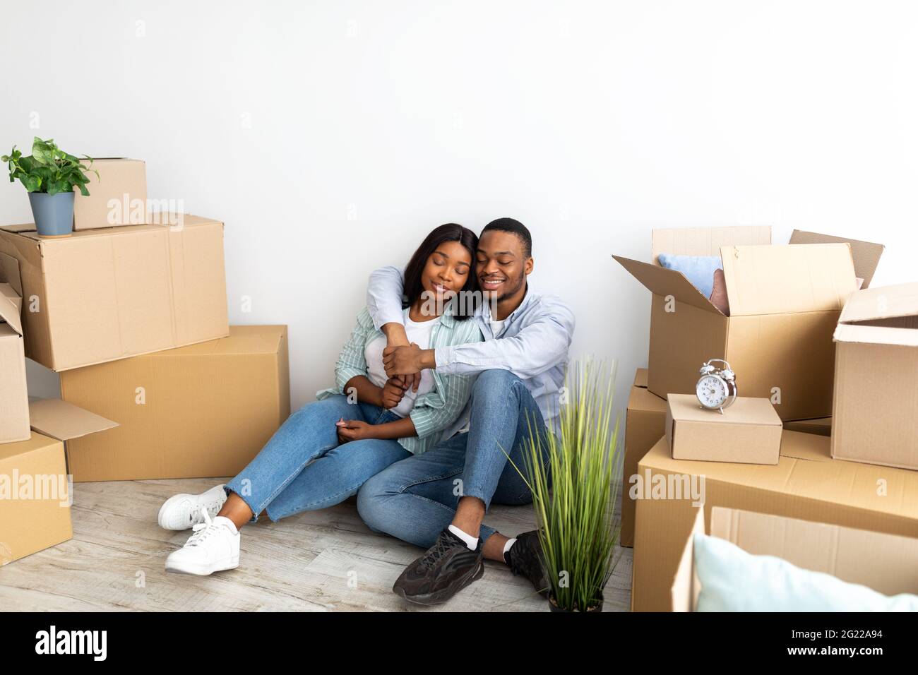 I coniugi neri amorevoli che si siedono fra le scatole mobili disimballate in nuova casa, abbracciando ed essendo felici di muoversi la propria casa Foto Stock