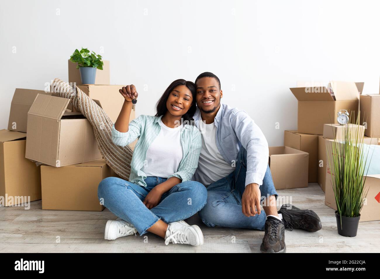 Felici sposi afroamericani con chiave della casa e scatole di cartone guardando la macchina fotografica e sorridendo, seduto sul pavimento Foto Stock