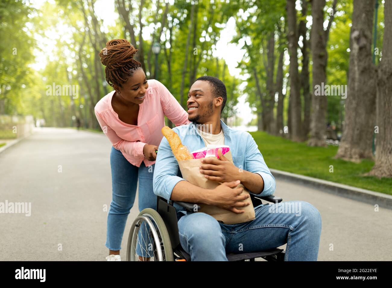 Felice giovane uomo disabile in sedia a rotelle trasportando borsa di carta con cibo, sua bella moglie aiutandolo all'aperto Foto Stock