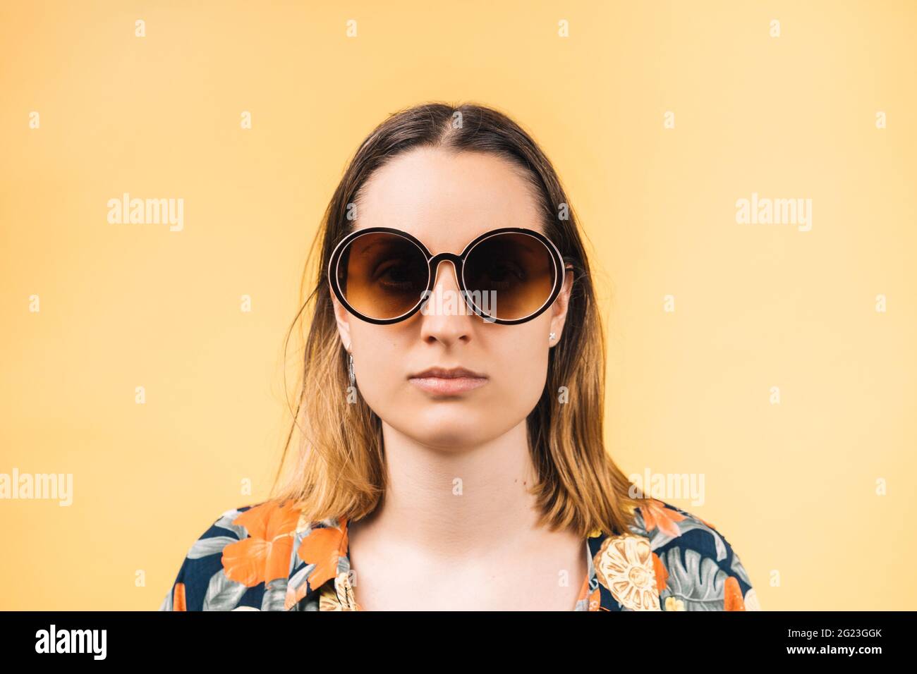 donna giovane seria che indossa una camicia fiorita e grandi occhiali da  sole rotondi con uno sfondo giallo Foto stock - Alamy