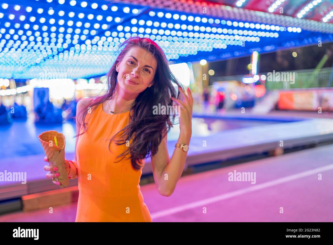 Gioiosa giovane donna vestita con un abito arancione che si posa di fronte a autoscontri al parco divertimenti, tenendo gelato Foto Stock