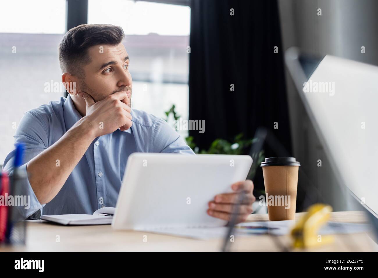 Uomo d'affari premuroso che tiene il tablet digitale vicino a un notebook e a un caffè sfocati Foto Stock