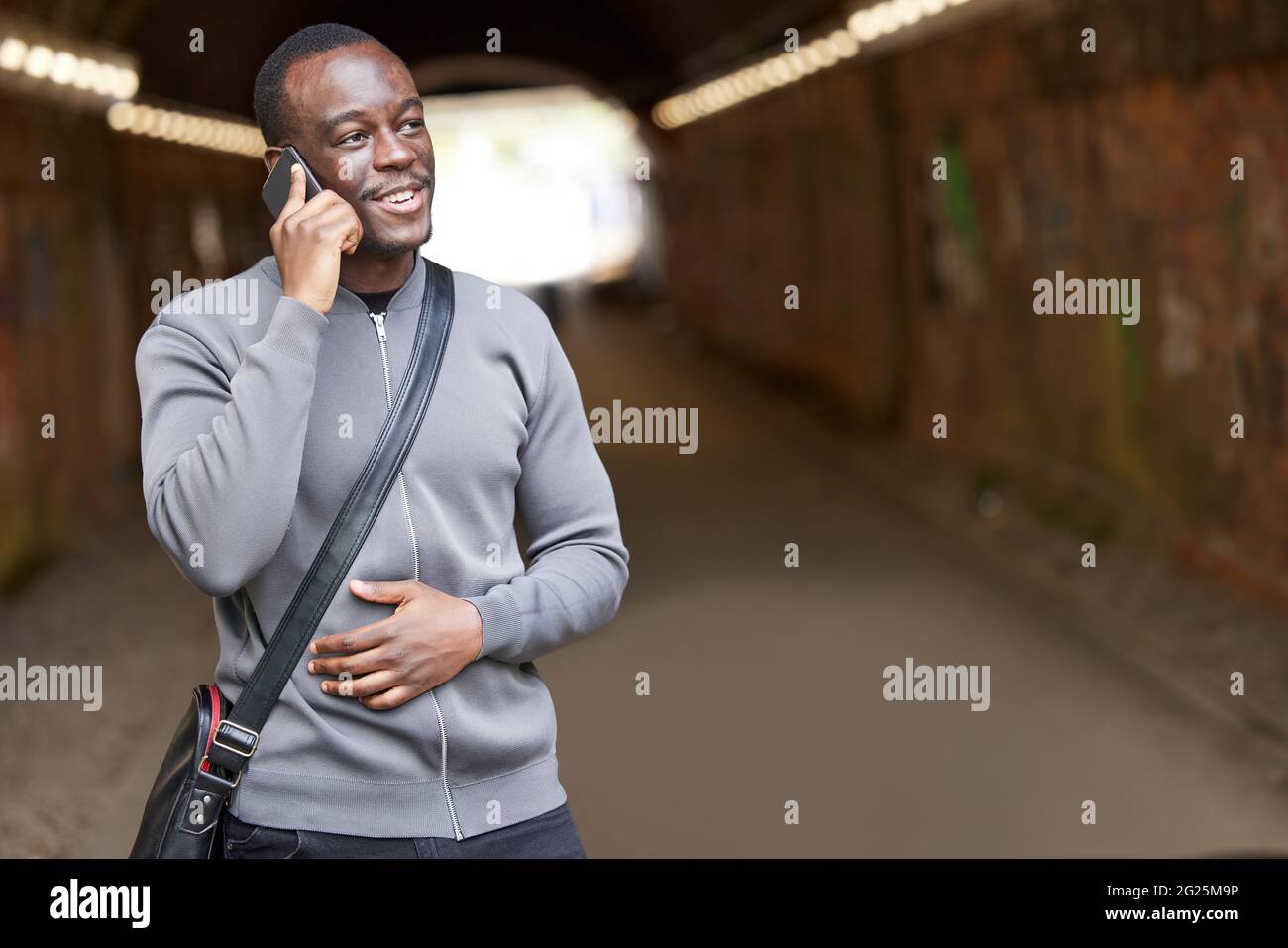 Sorridente giovane uomo in ambiente urbano in piedi in Tunnel che parla sul telefono cellulare Foto Stock