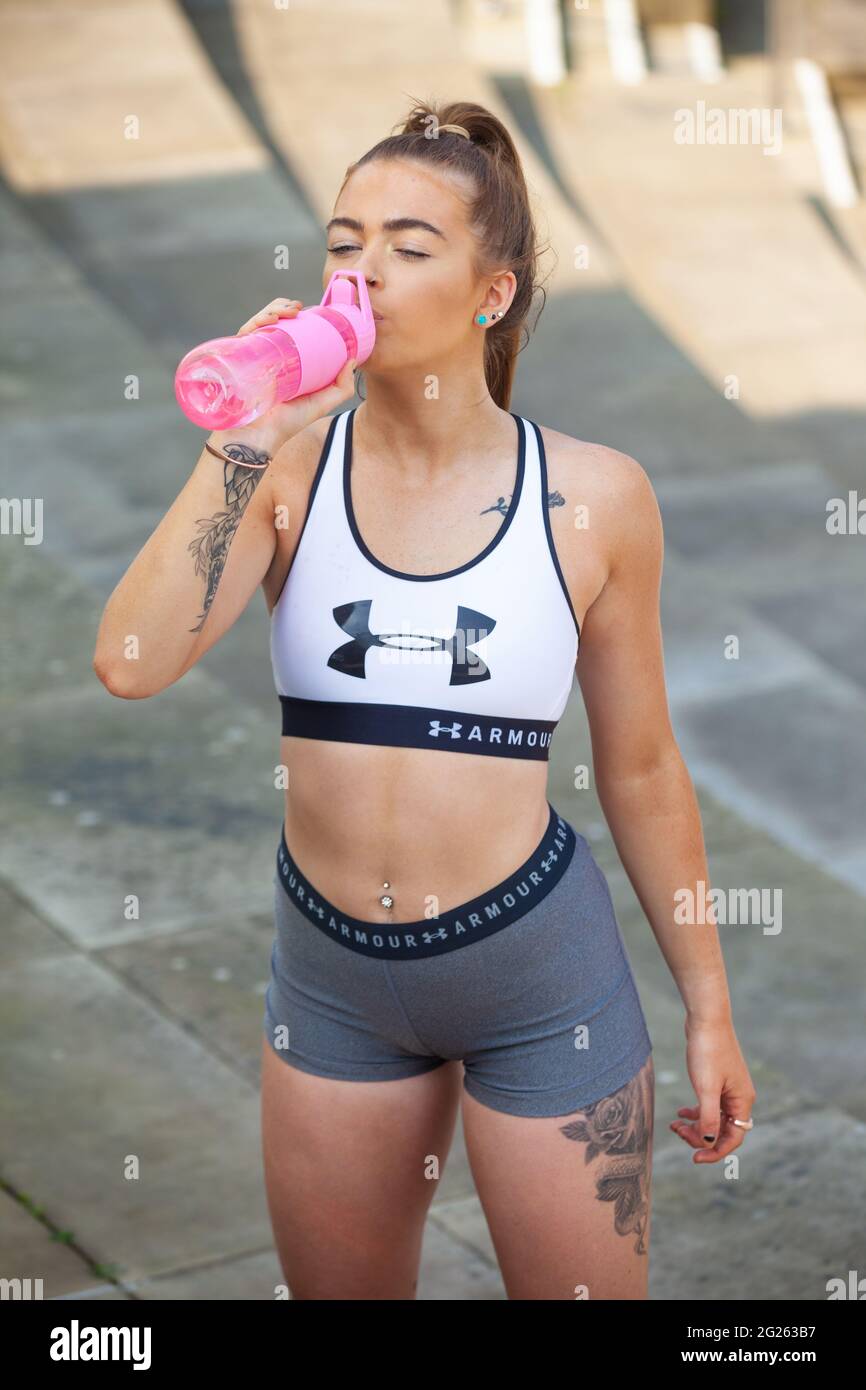 Una giovane donna che indossa abiti sportivi all'aperto e che beve da una bottiglia d'acqua rosa. Foto Stock