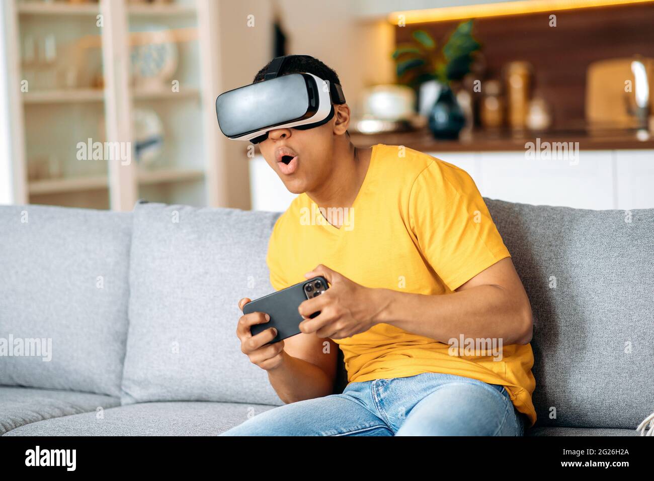 Eccitato ragazzo di razza mista latino seduto su un divano a casa in abiti casual, utilizza un dispositivo di realtà virtuale, in un mondo digitale, gesturing con le mani, tiene smartphone. Giocare nel mondo virtuale Foto Stock