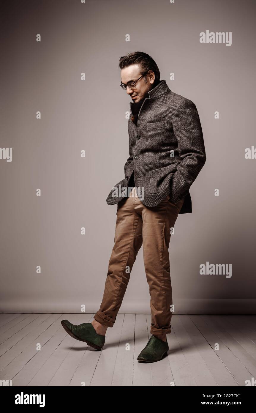 Uomo adulto sorridente in giacca a scacchi e sciarpa nera si alza con le mani in tasca, guardando giù la sua scarpa Foto Stock