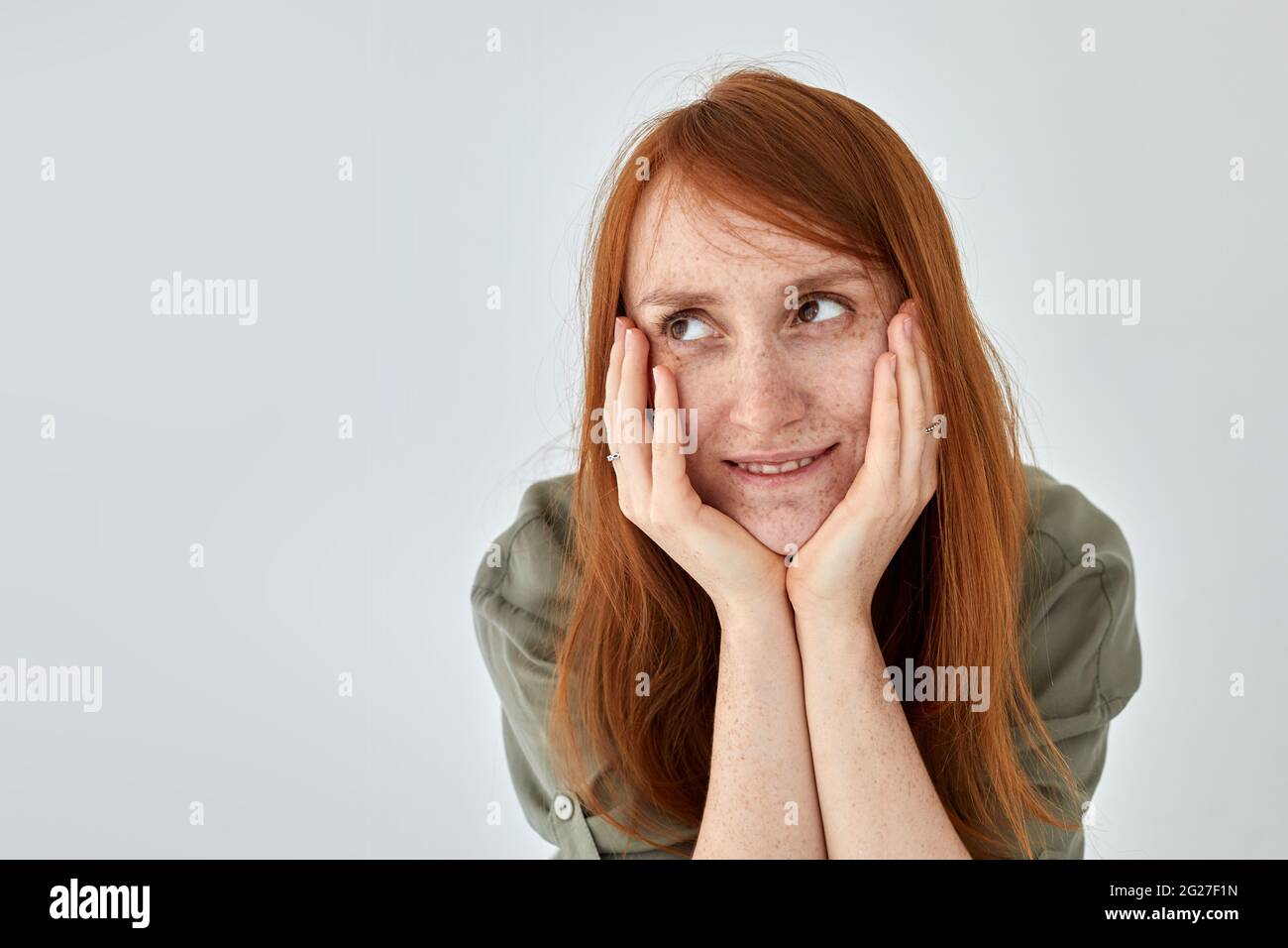 Donna positiva con capelli di zenzero che tiene le mani sulle guance e che guarda via contro sfondo bianco Foto Stock