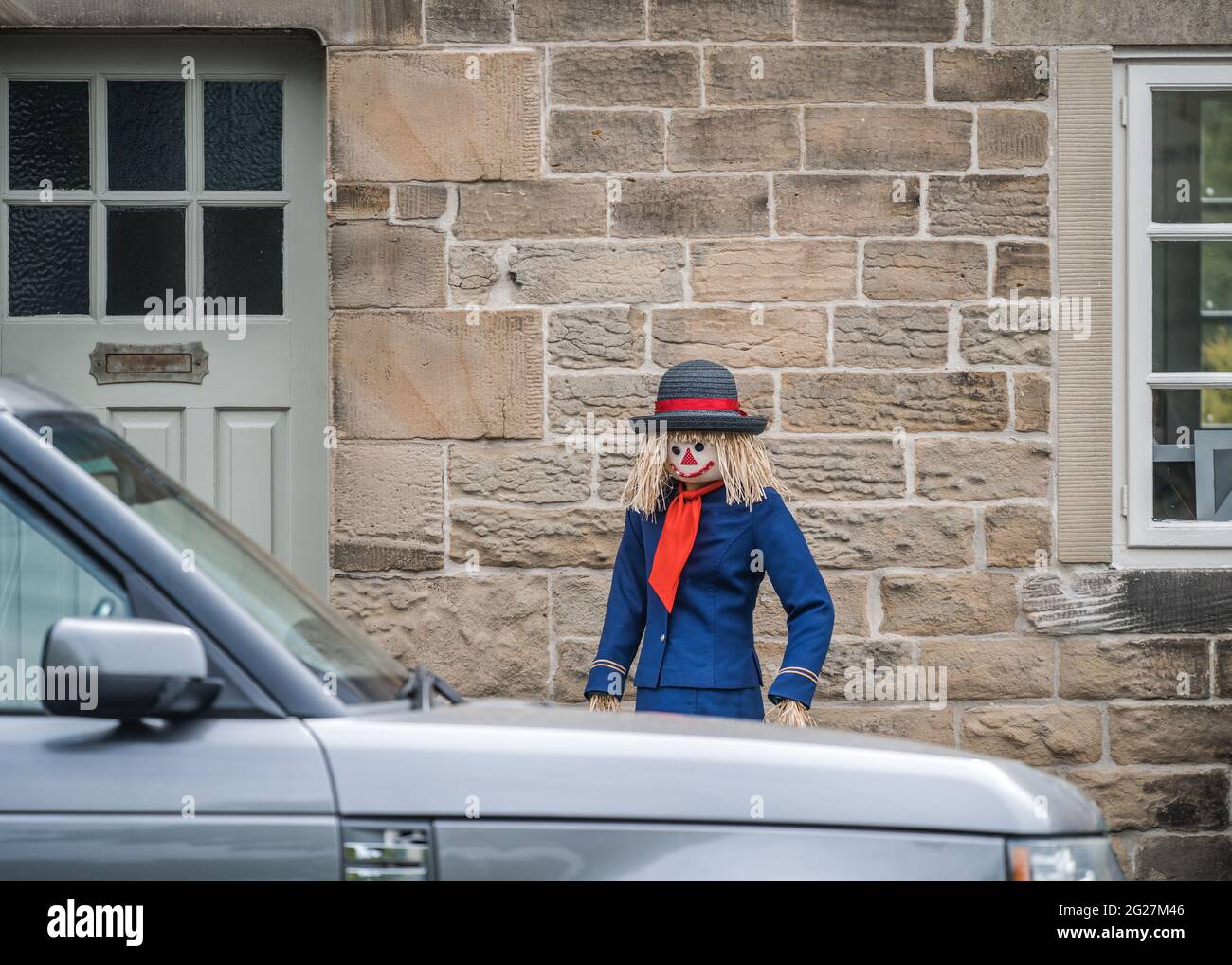 Scarecrow donna assistente di volo si trovava fuori vecchio cottage in pietra in campagna. Vestiti in uniforme blu con cappello dell'equipaggio e pappagallo di paglia Foto Stock