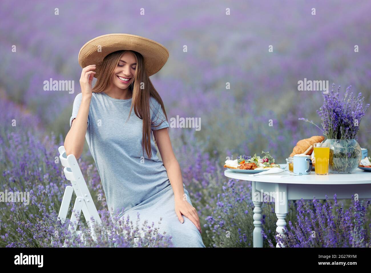 Vista frontale della giovane donna seduta al tavolo con croissant, succo  d'arancia e cibo. Bella ragazza che indossa cappello di paglia e abito  lungo seduto in campo di lavanda e sorridente, ristorante