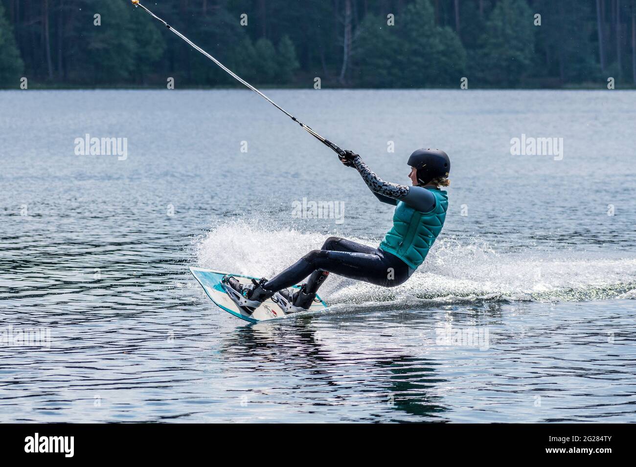 Ragazza con casco e giubbotto di salvataggio fa un wakeboard sul lago con foresta sullo sfondo, wakeboarding, sport acquatici in estate Foto Stock
