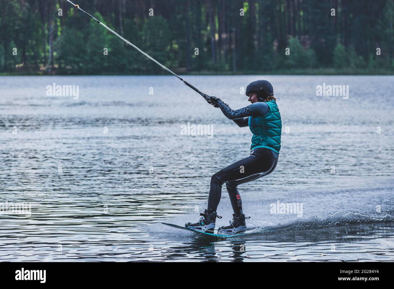 Ragazza con casco e giubbotto di salvataggio fa un wakeboard sul lago con foresta sullo sfondo, wakeboarding, sport acquatici in estate Foto Stock