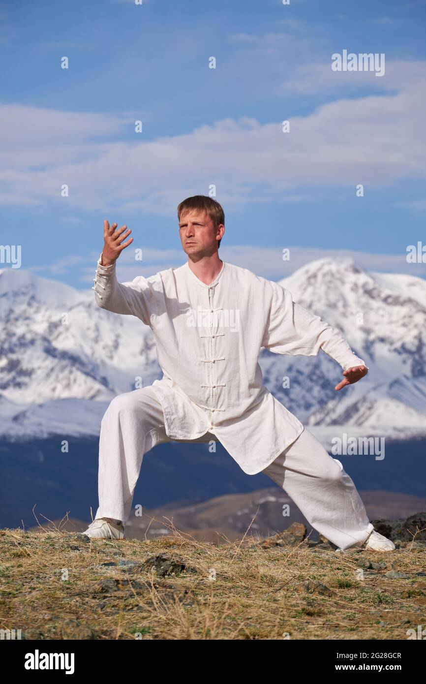 Wushu master in una formazione uniforme sportiva bianca sulla collina. Il campione di Kungfu allena le arti maritiche nella natura sullo sfondo delle montagne innevate. Foto Stock