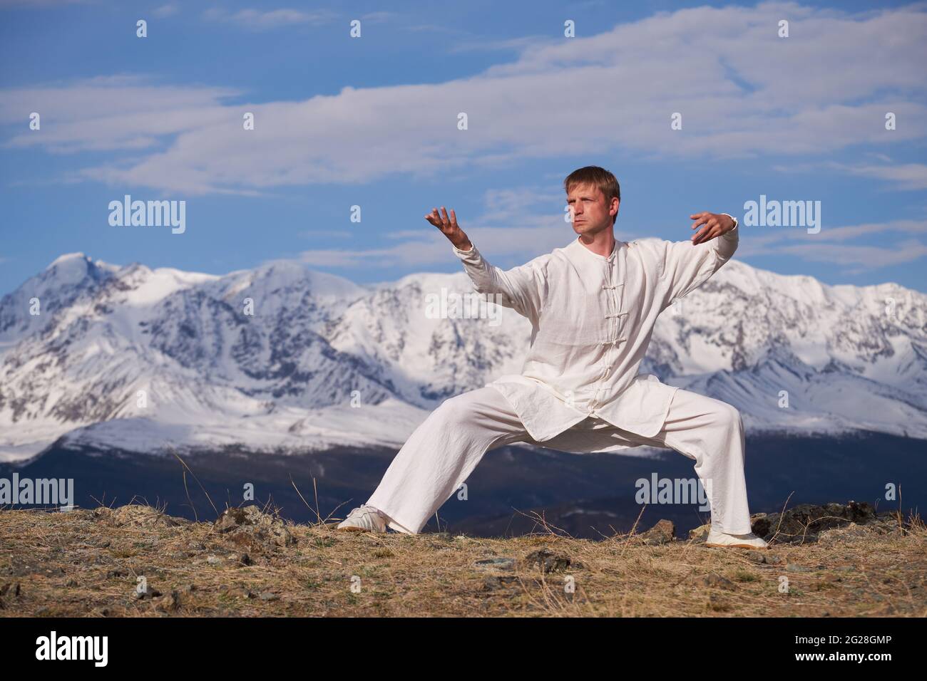 Wushu master in una formazione uniforme sportiva bianca sulla collina. Il campione di Kungfu allena le arti maritiche nella natura sullo sfondo delle montagne innevate. Foto Stock