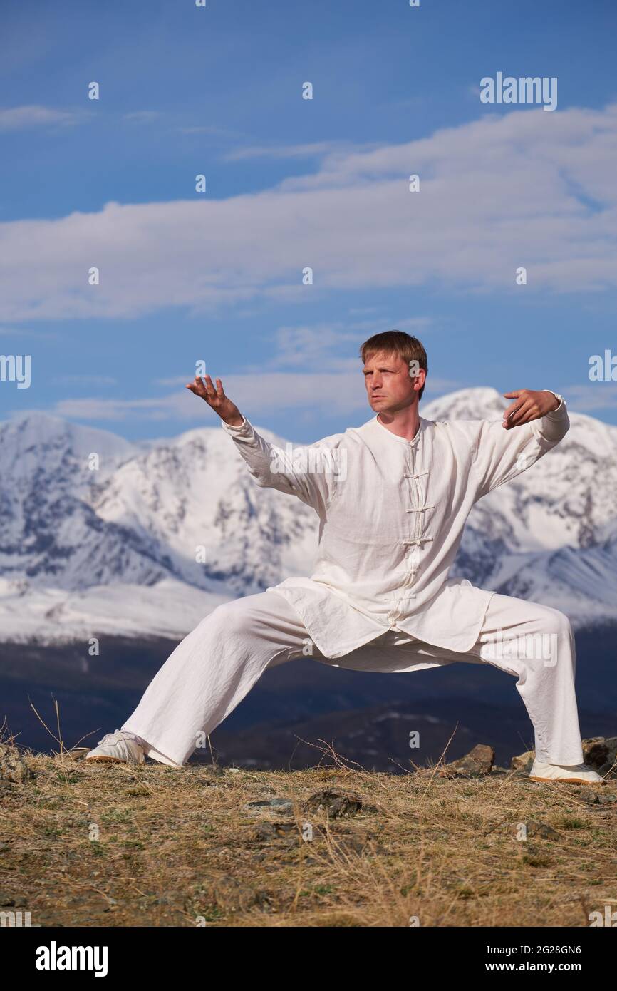 Wushu master in una formazione uniforme sportiva bianca sulla collina. Il campione di Kungfu allena le arti maritiche nella natura sullo sfondo delle montagne innevate. Foto Stock