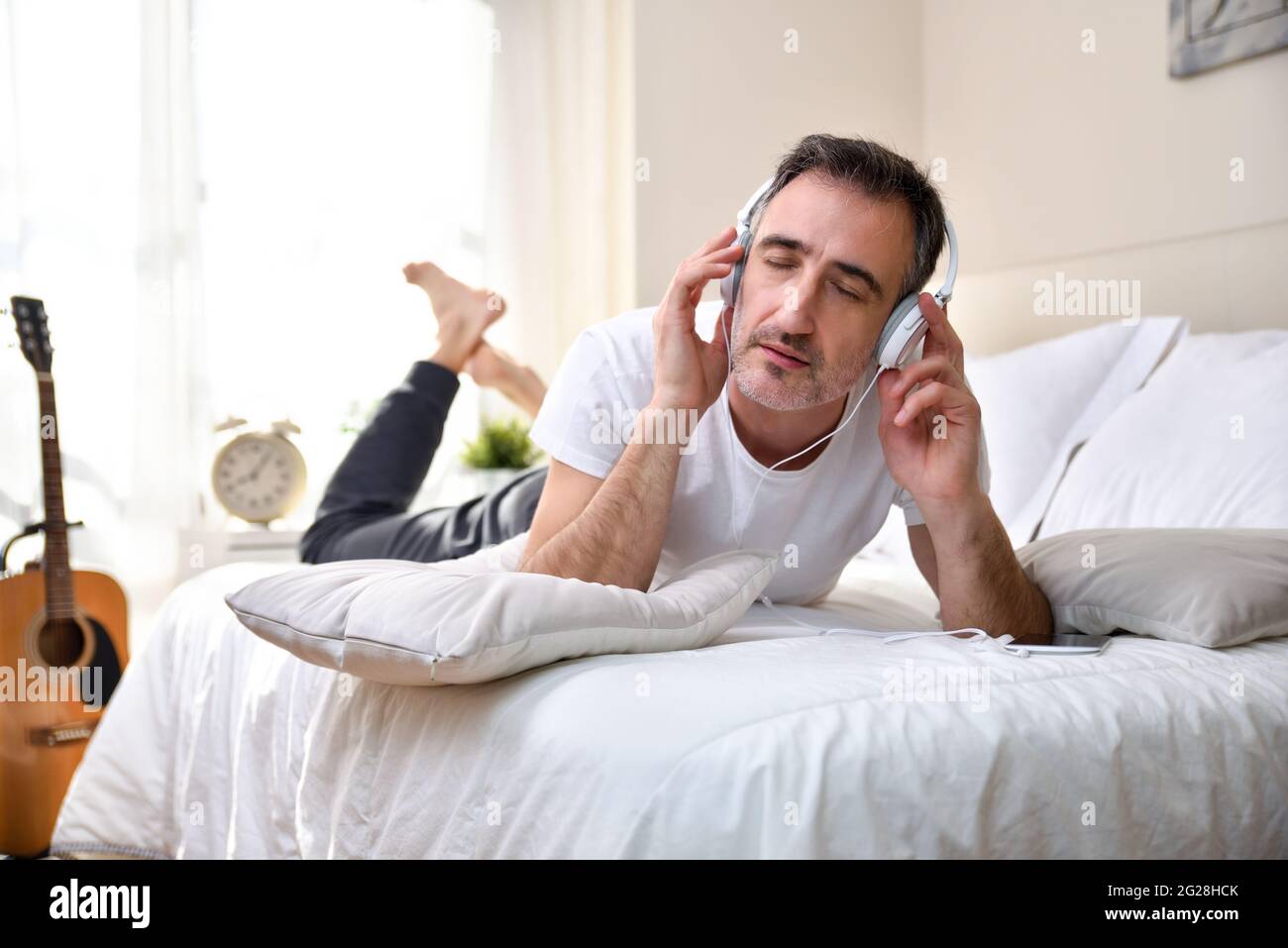 L'amante della musica ascoltò concentrato sdraiato sul letto da un cellulare con cuffie bianche con chitarra e finestra sullo sfondo che entra alla luce del sole Foto Stock