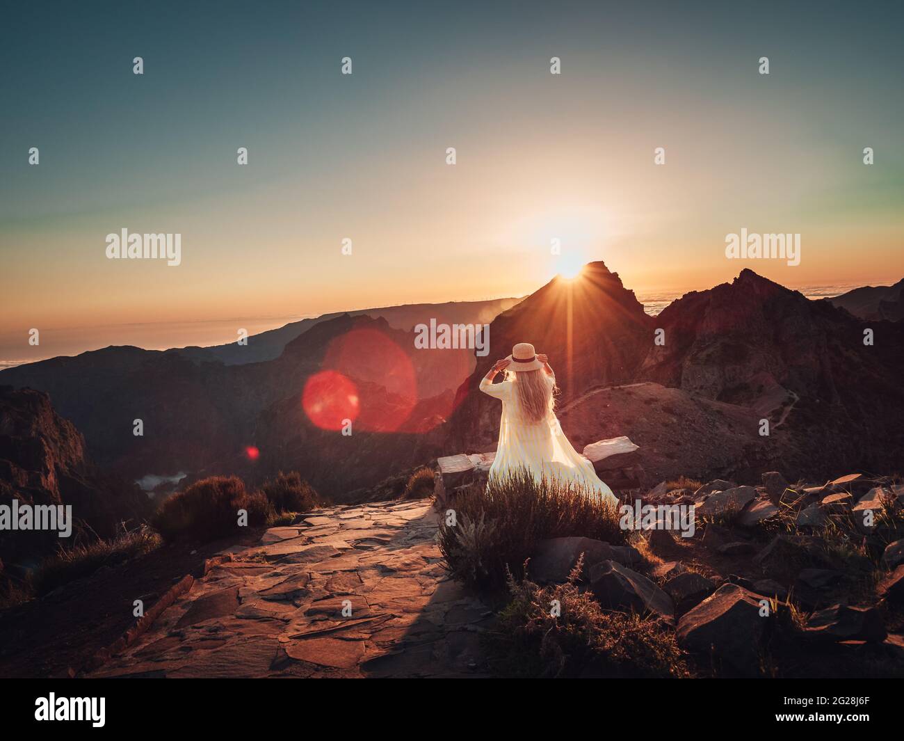 Donna dalla schiena in un abito bianco e cappello in una zona rocciosa al tramonto Foto Stock