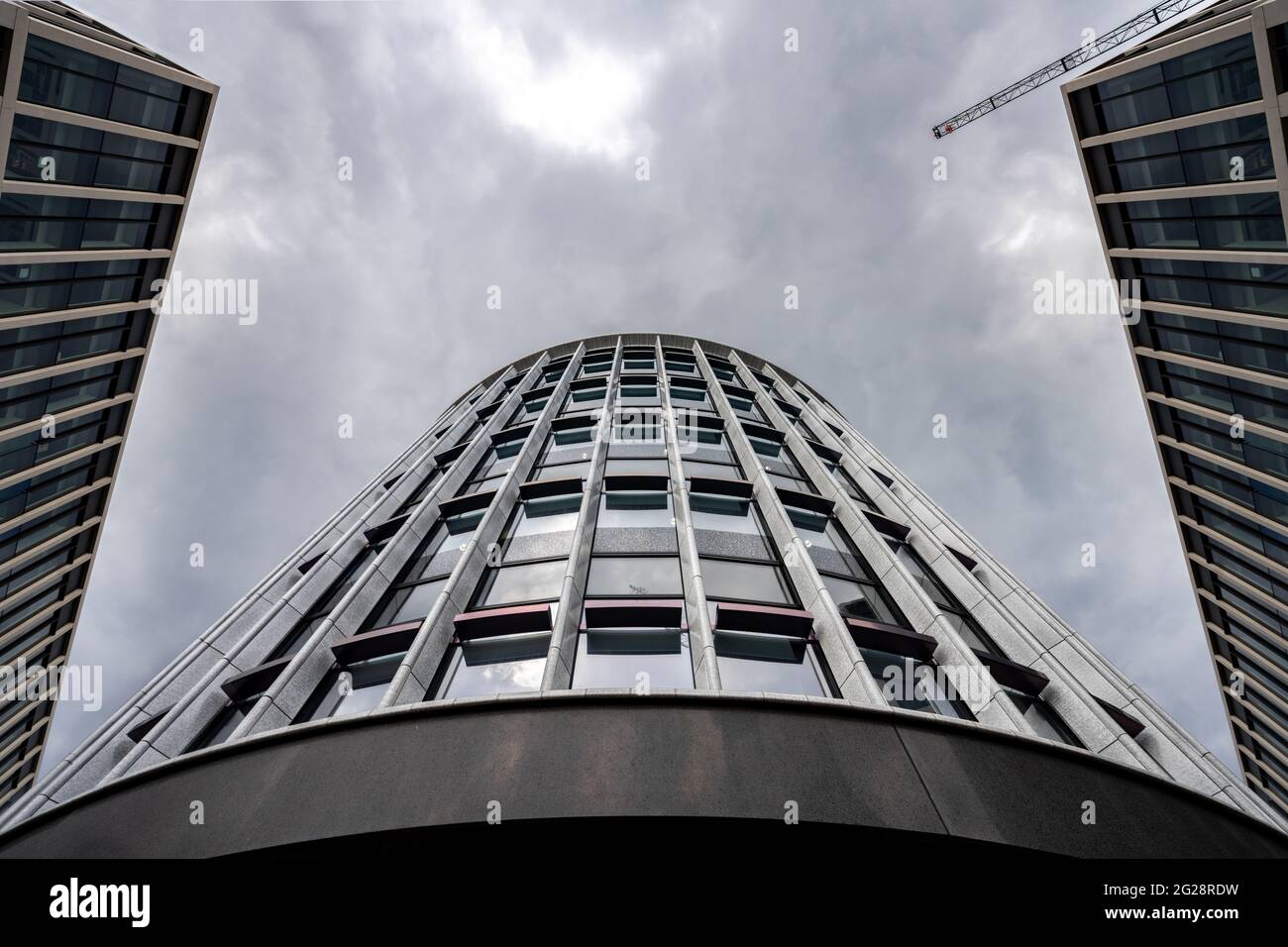 Moderno edificio futuristico che guarda sullo skyline con edifici simmetrici e gru a torre in costruzione di alto livello. Blocco da ufficio con curva Foto Stock