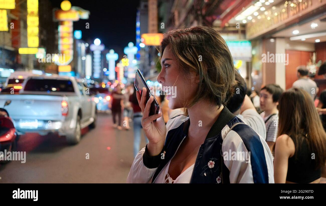 Giovane Asian Lady capelli scuri parlando su un cellulare Chinatown Bangkok Thailandia offuscato fuori sfondo luci al neon gente di notte in background Foto Stock