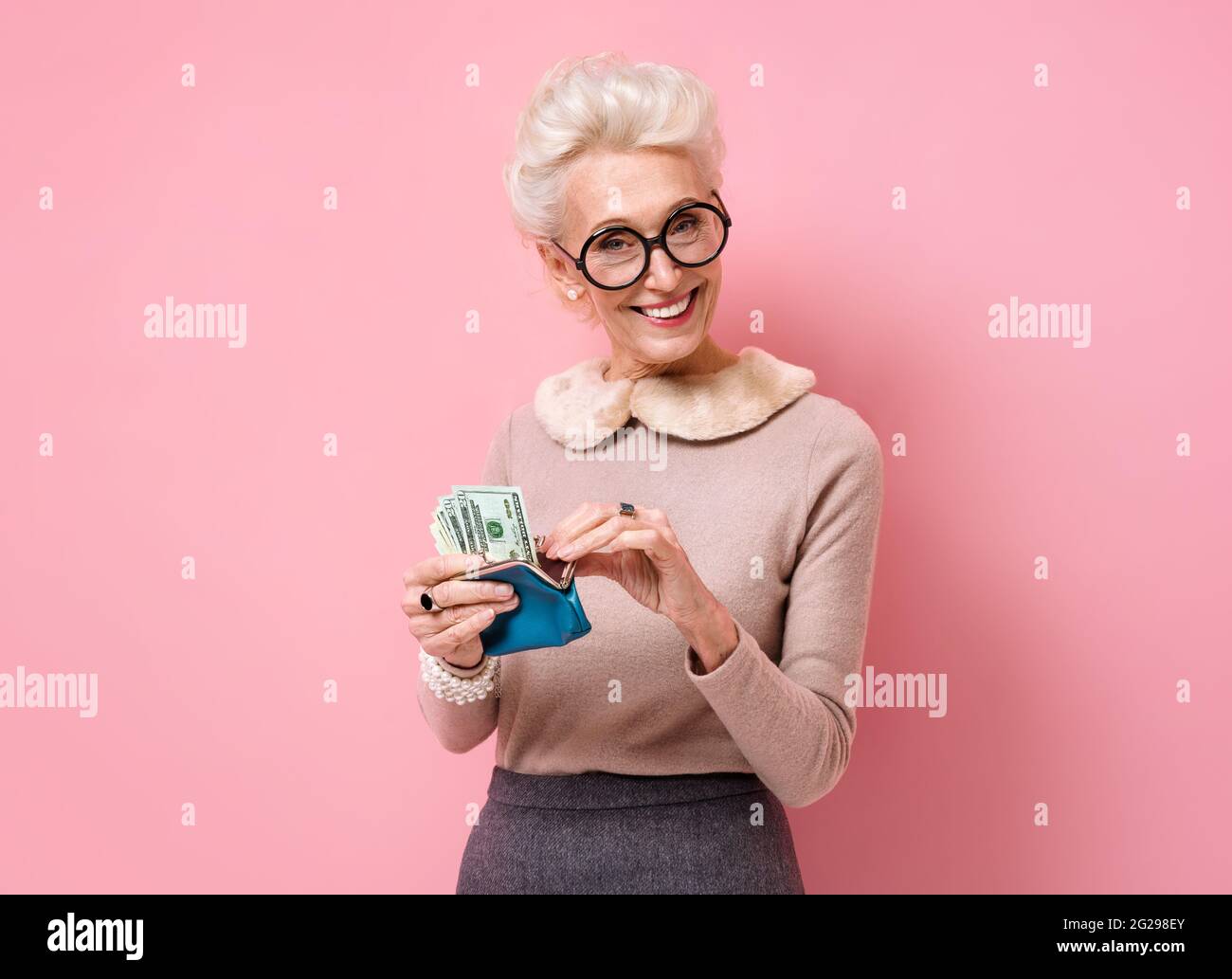 Donna sorridente che guarda il suo portafoglio. Foto di donna anziana gentile indossa occhiali su sfondo rosa. Foto Stock