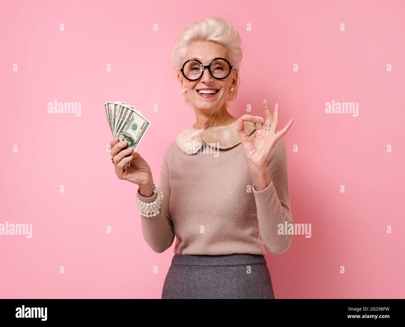 La nonna sorridente mostra denaro in contanti e fa un gesto corretto, dimostra che tutto va bene. Foto di donna anziana gentile indossa occhiali sul pin Foto Stock