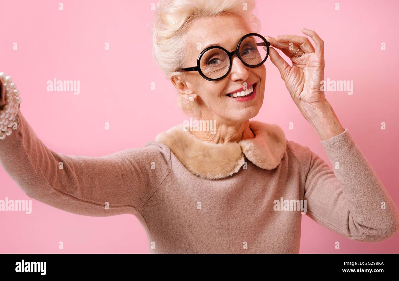 Nonna allegra che prende selfie al telefono. Foto di donna anziana gentile indossa occhiali su sfondo rosa. Foto Stock