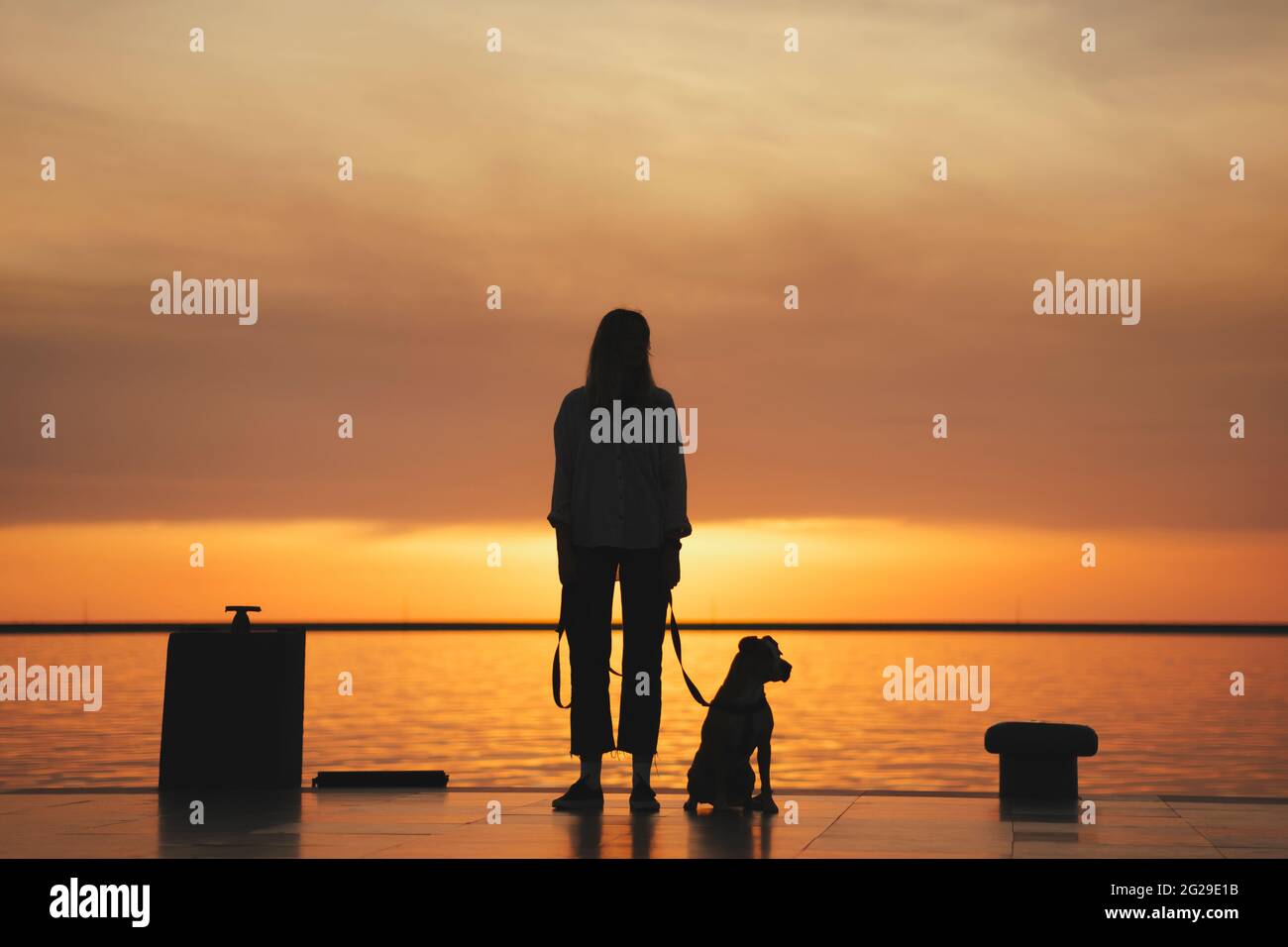 Silhouette di una donna con cane al molo vicino all'acqua, ora d'oro Foto Stock