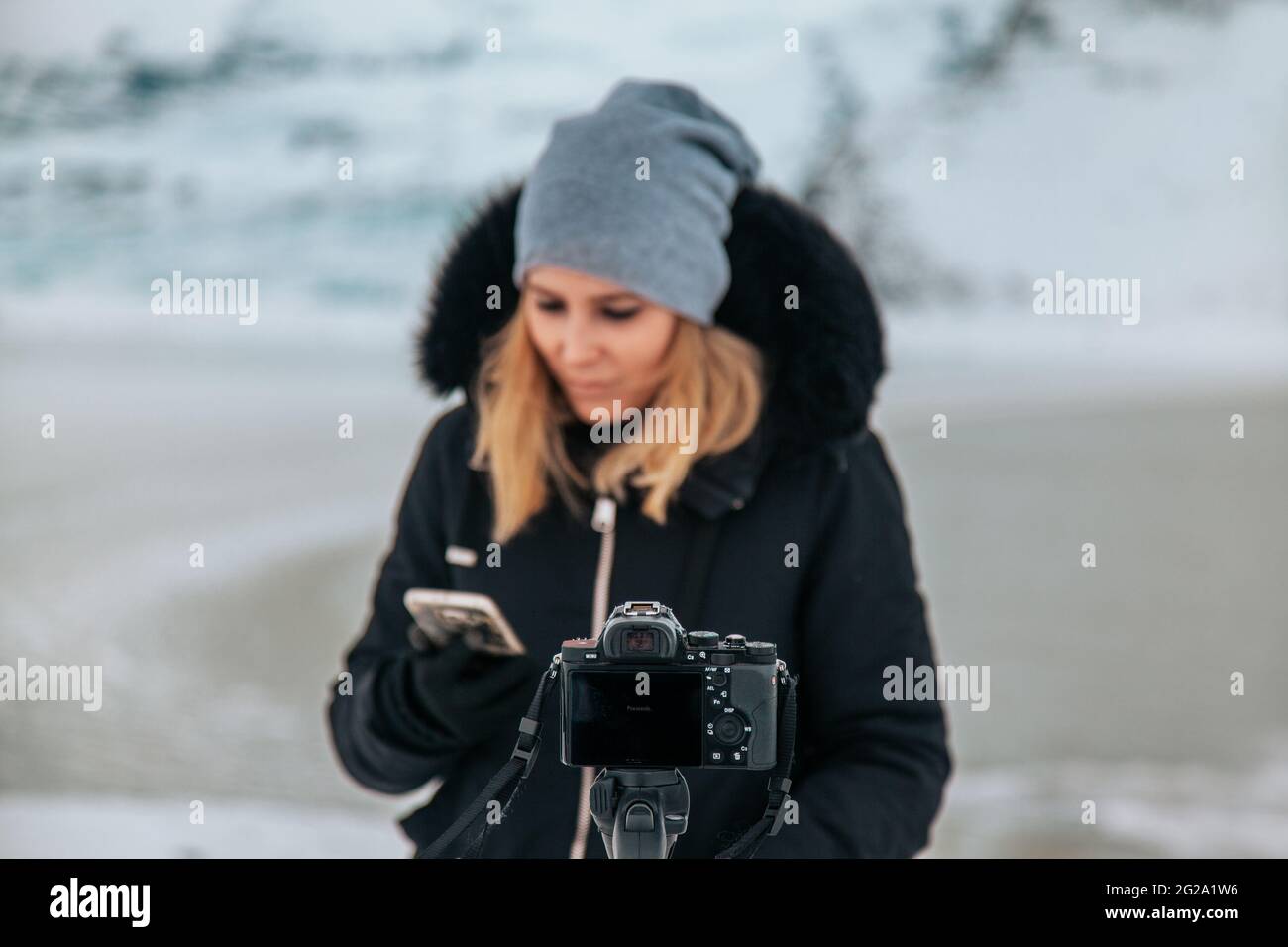 Giovane viaggiatore femminile con capelli biondi in abiti caldi e cappello in piedi davanti alla fotocamera moderna con treppiede tra le montagne ghiacciate in Islanda utilizzando il cellulare Foto Stock