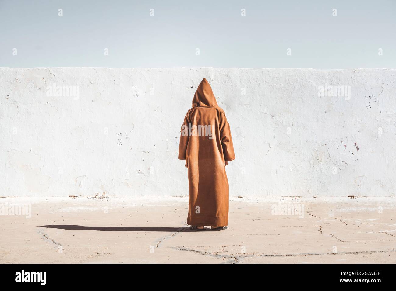 Vista posteriore di una persona in un accappatoio con cappuccio marrone che si trova vicino a una parete bianca in un campo di rifugio nel deserto del Sahara Foto Stock