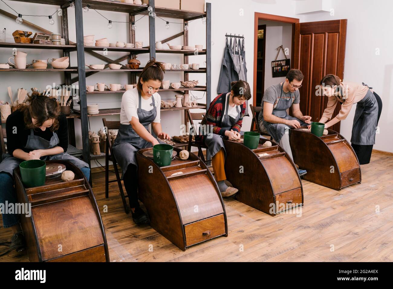 Persone entusiaste in grembiuli che lavorano con la scultura in argilla su ruota in ceramica con l'aiuto di artigiani in grande officina leggera Foto Stock