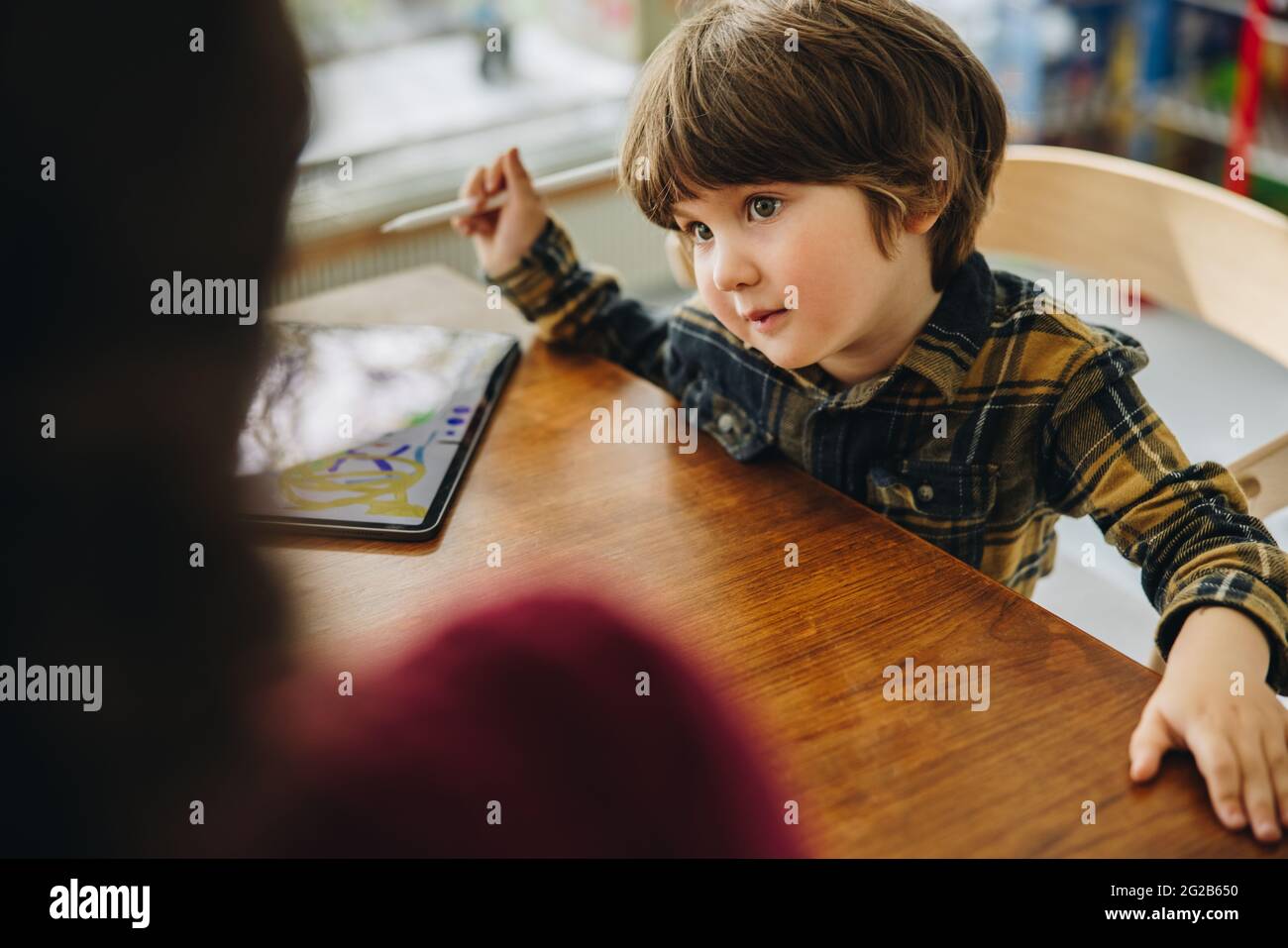 Ragazzo innocente che guarda la mamma mentre si siede a tavola con un tablet digitale. Figlio carino che guarda la madre mentre disegnano su un tablet pc. Foto Stock