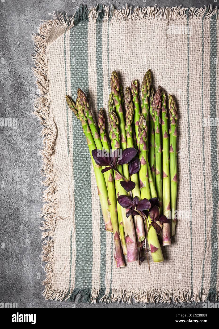 Vista dall'alto di un fascio di asparagi verdi freschi con foglie di basilico viola su una tovaglia dalla linea rustica. Foto Stock
