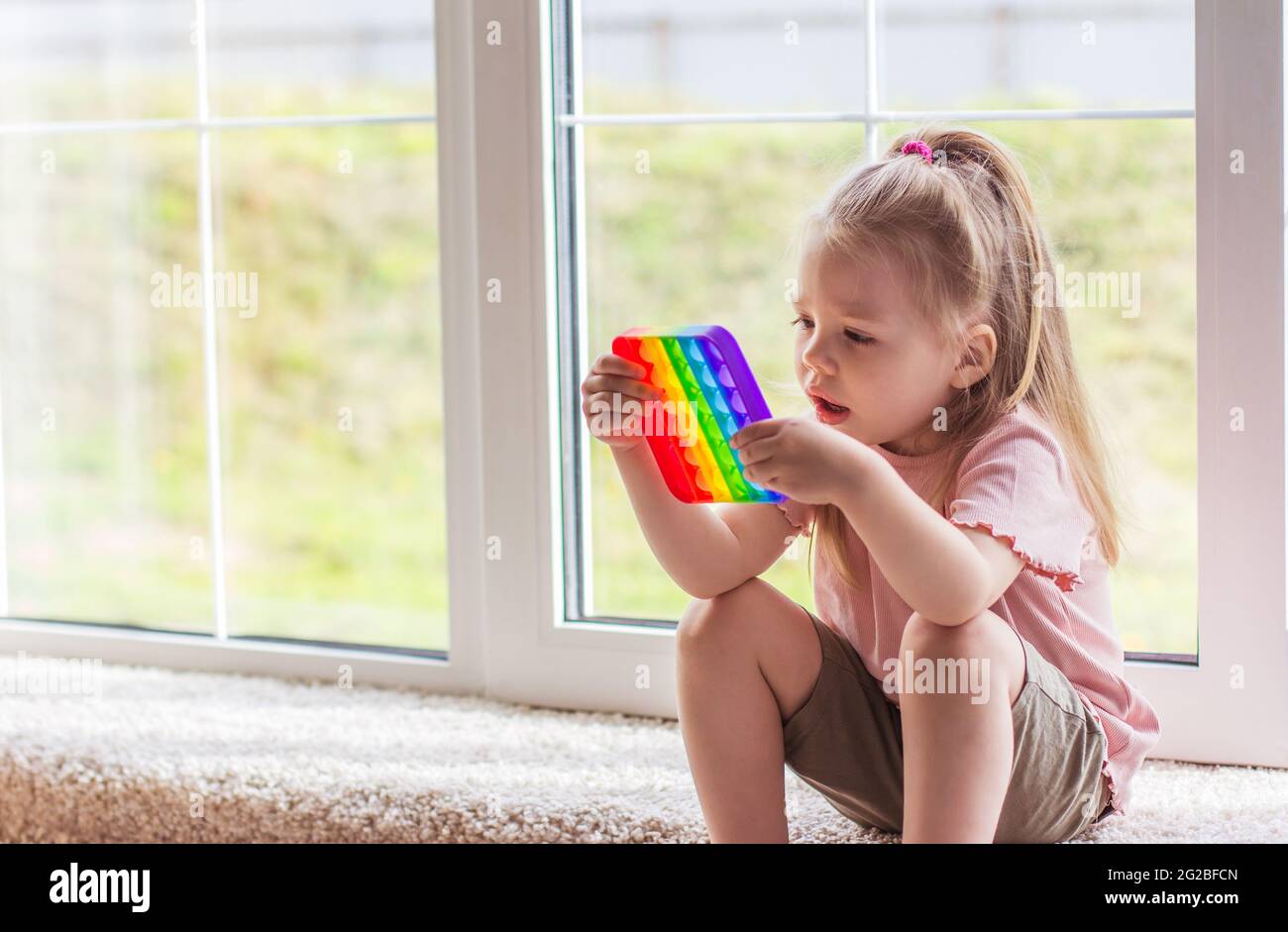 I bambini della bambina bionda giocano con il nuovo giocattolo sensoriale di tendenza - arcobaleno pop esso. Giocattolo antistress per bambini e adult.colorful giocattolo semplice concavità. Squis Foto Stock