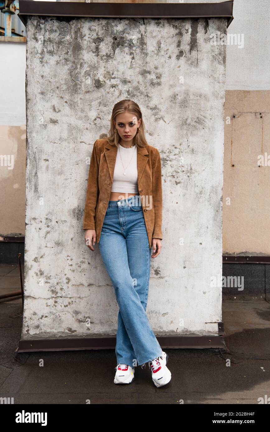 modello completo in jeans, blazer in pelle scamosciata ed eleganti sneaker  in posa con gambe incrociate vicino alla parete in cemento sul tetto Foto  stock - Alamy