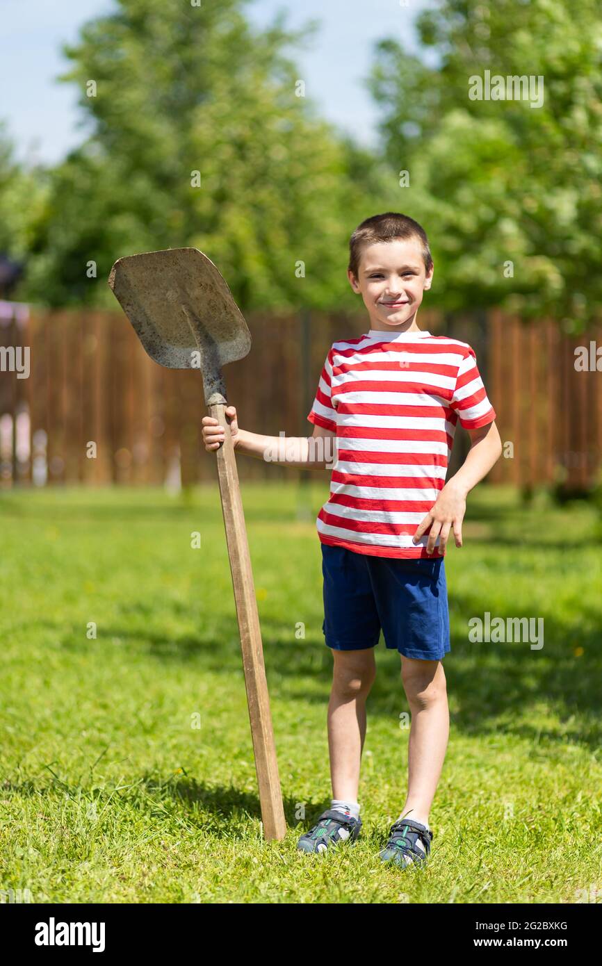 Un ragazzino allegro si alza e tiene in mano una pala, pronta a lavorare nel giardino di una casa di campagna. Foto Stock