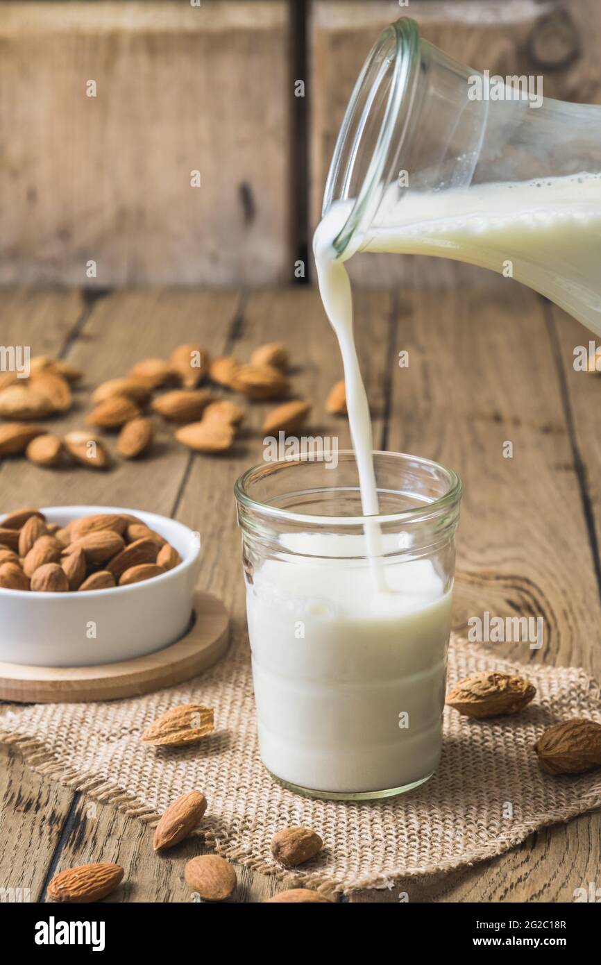 Latte di mandorle vegane versato in un bicchiere, noccioli di mandorle e mandorle intere sul vecchio tavolo rustico in legno, verticale Foto Stock