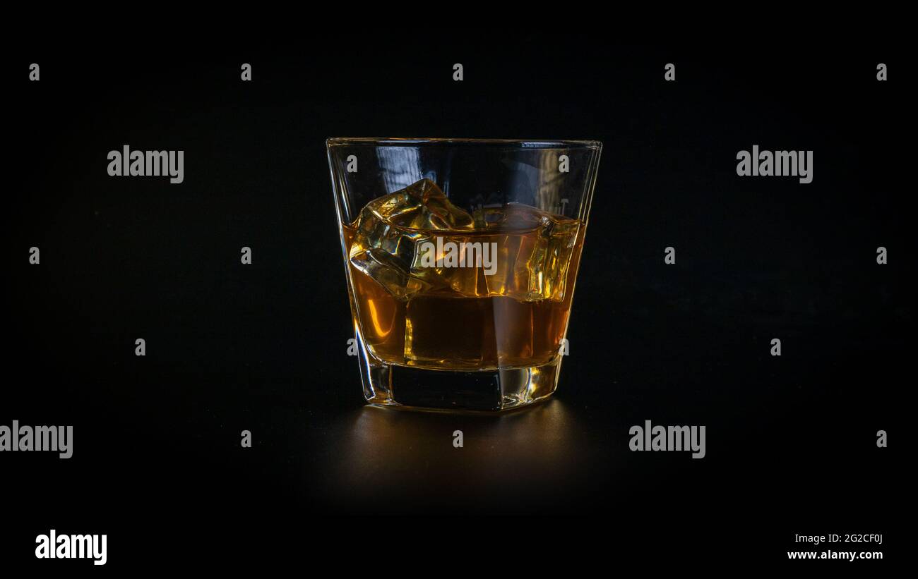 Bevanda o cocktail di whisky, marrone miele, in un bicchiere, pieno di cubetti di ghiaccio freddo e frizzante. Sfondo nero distinto, riflesso di vetro sullo sfondo. Foto Stock