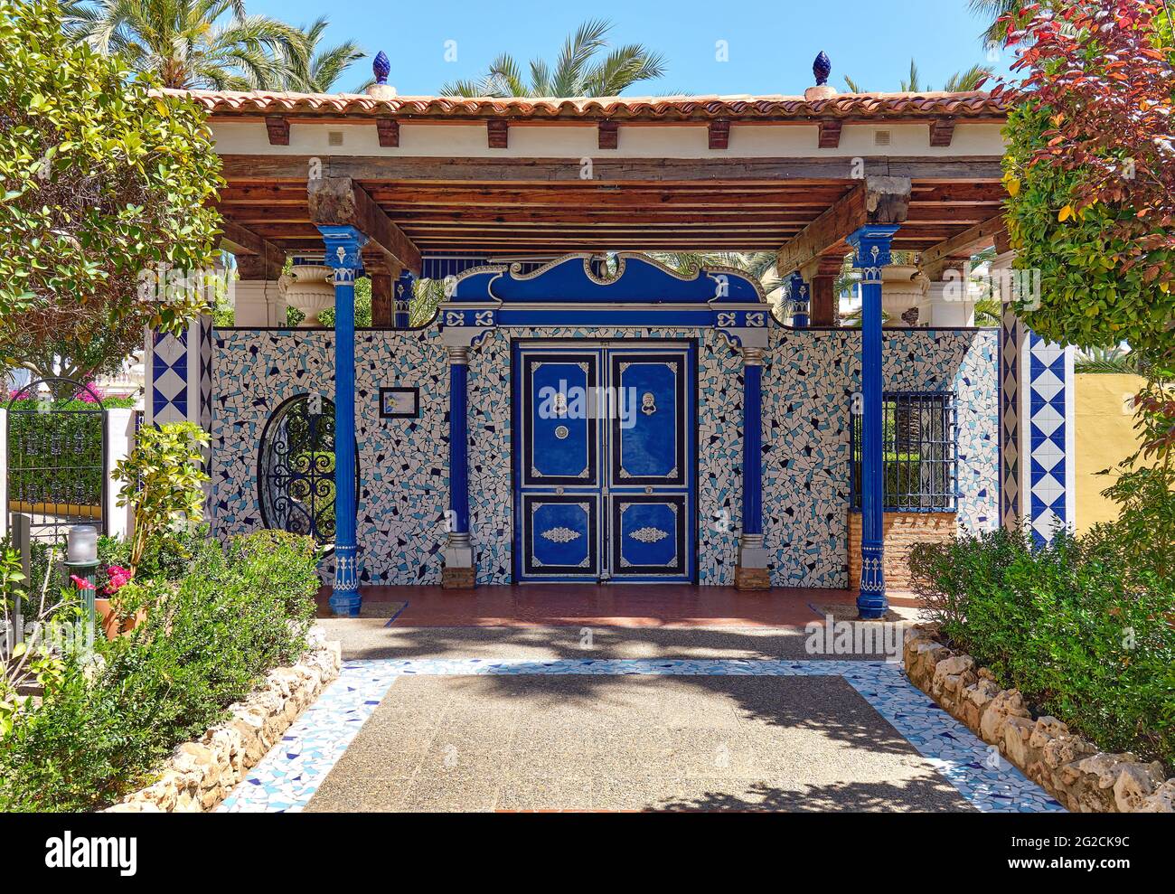 Percorso fiancheggiato da piante verdi rigogliose che conduce a un ingresso decorativo classico con piastrelle in ceramica azulejo. Giorno estivo soleggiato, nessuna persona. Provincia di Alicante Foto Stock