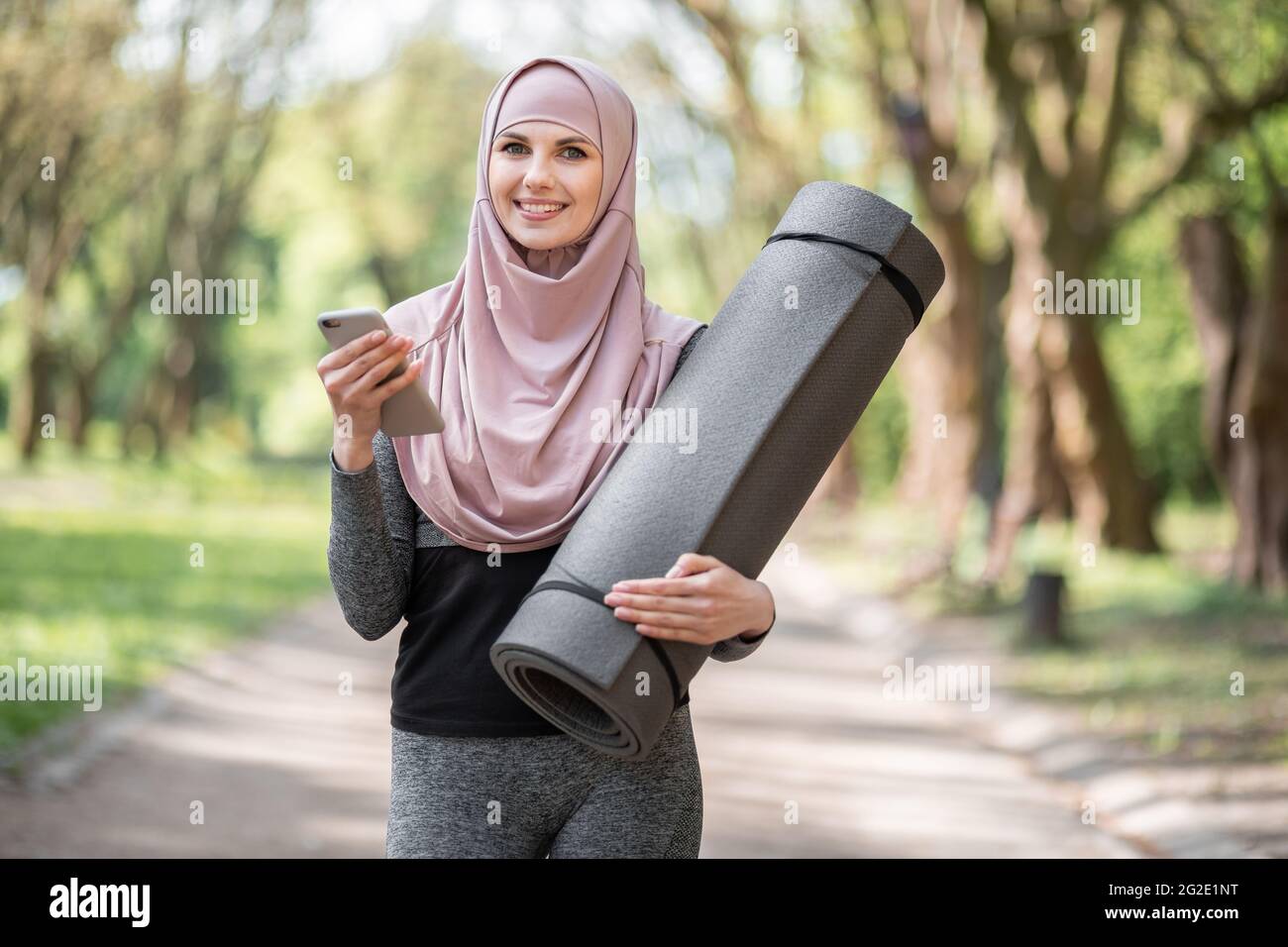 Vista frontale di una donna sorridente in hijab e outfit sportivo utilizzando un moderno telefono cellulare mentre si prende una pausa durante l'allenamento all'aperto. Concetto di stile di vita attivo. Foto Stock
