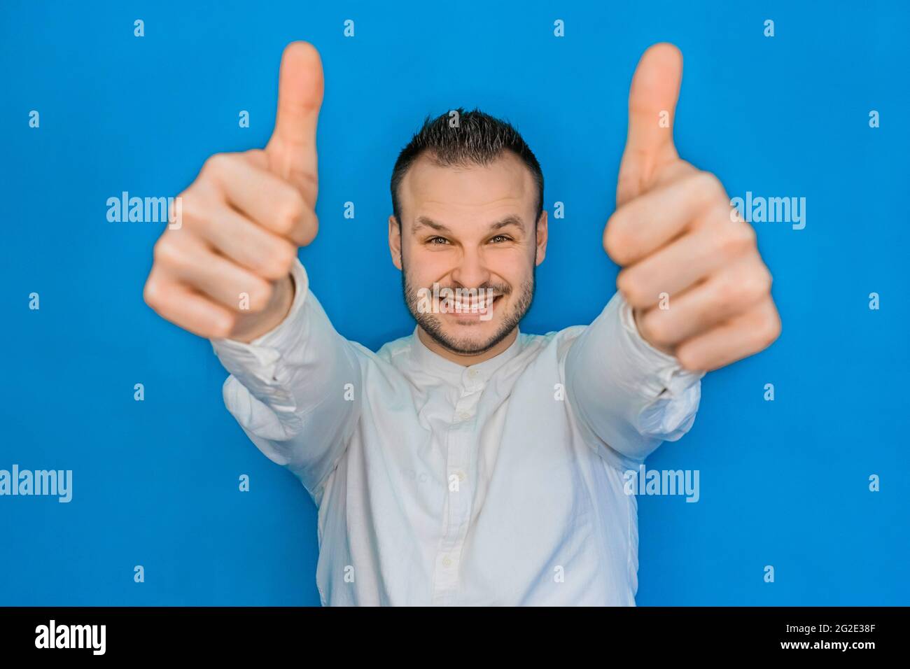 Ritratto di giovane attraente uomo d'affari felice europeo in camicia bianca con barba che mostra i pollici cool su due mani su sfondo blu. Foto Stock