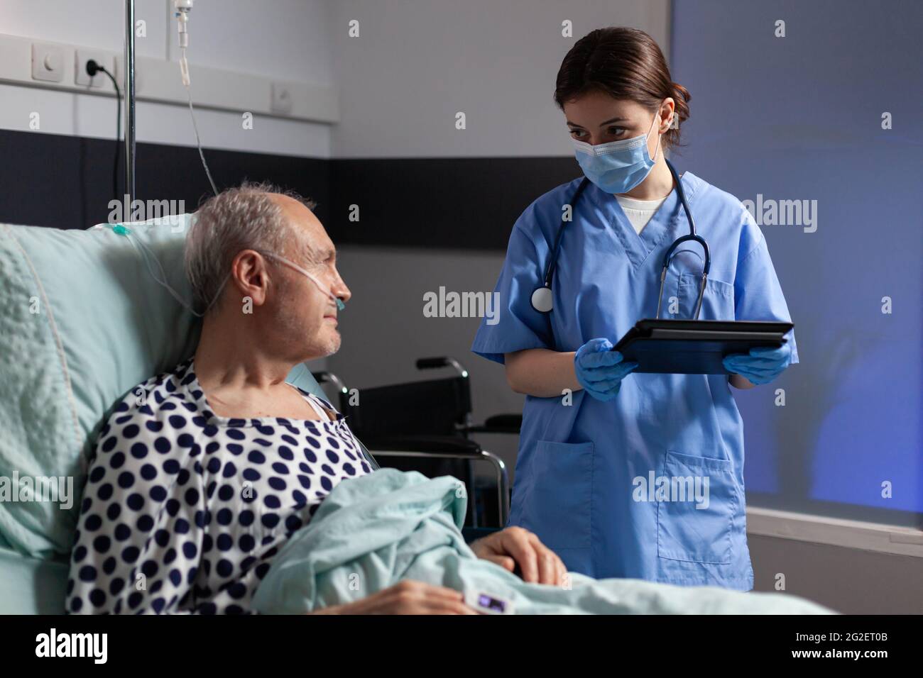 Assistente medico con maschera di protezione controllo trattamento di uomo anziano leggere note su tablet pc. Ammalato anziano che si trova a letto discutendo con l'infermiere circa l'esperienza di diagnosi con la sacca per infusione attaccata. Foto Stock