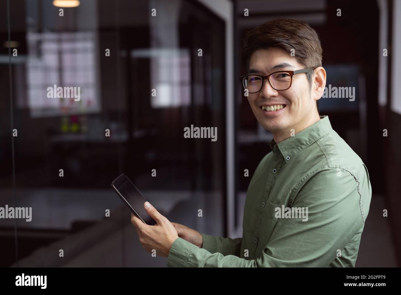 Ritratto di un uomo d'affari asiatico sorridente in piedi in un ufficio vuoto utilizzando un tablet Foto Stock