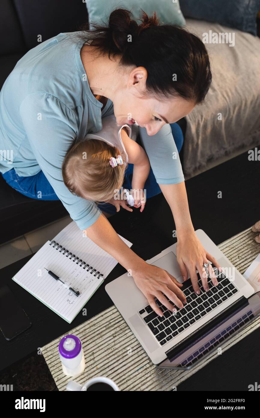Sorridente madre caucasica che tiene il suo bambino usando il laptop mentre lavora da casa Foto Stock