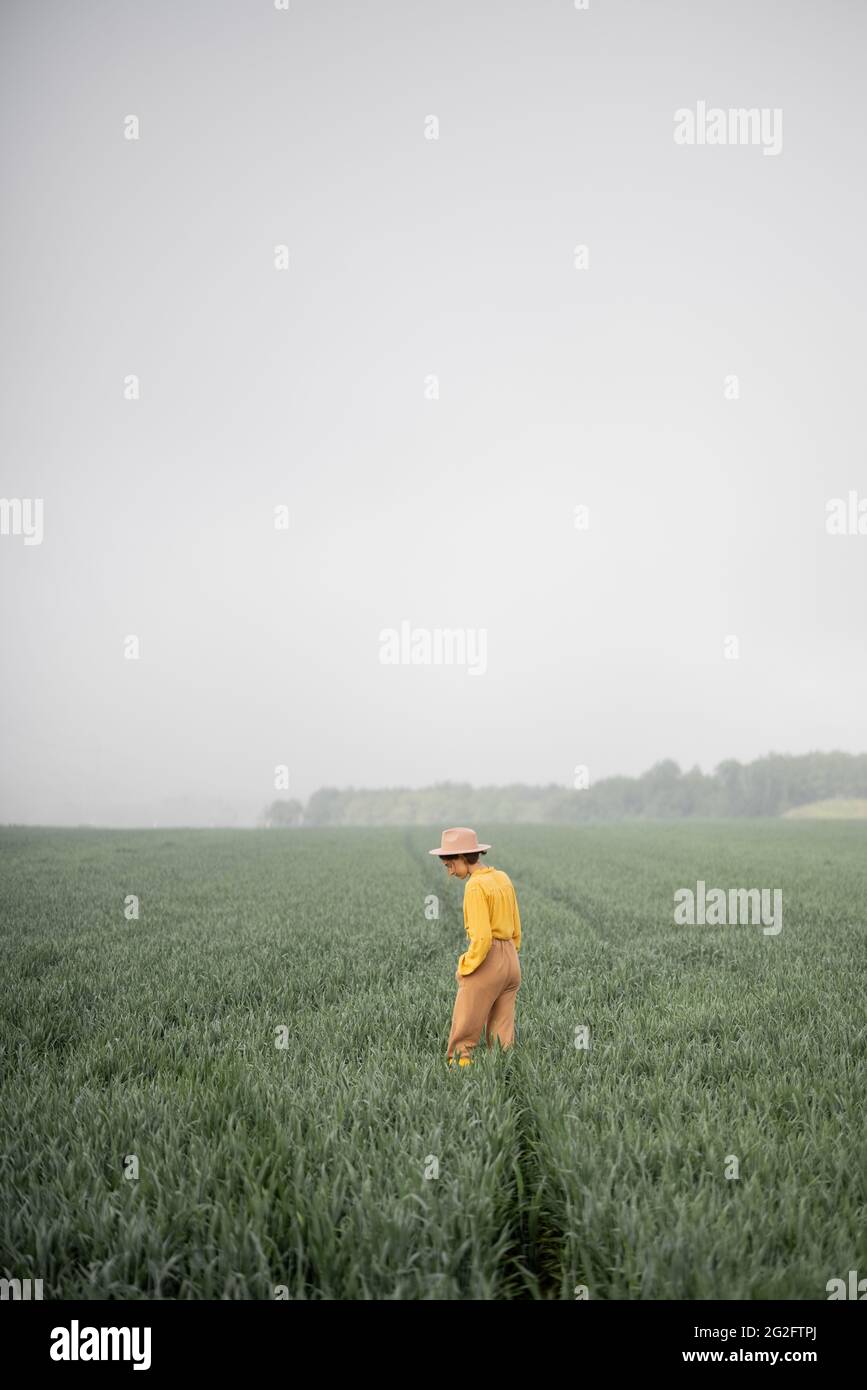 Donna in cappello che cammina sul campo di grano verde durante il tempo foggy. Campagna e vita tranquilla. Foto Stock