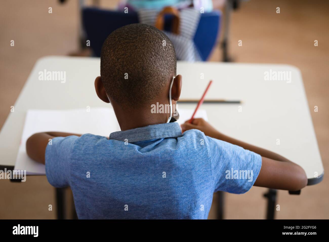 Vista posteriore del ragazzo afroamericano che studia mentre si siede sulla sua scrivania nella classe a scuola Foto Stock
