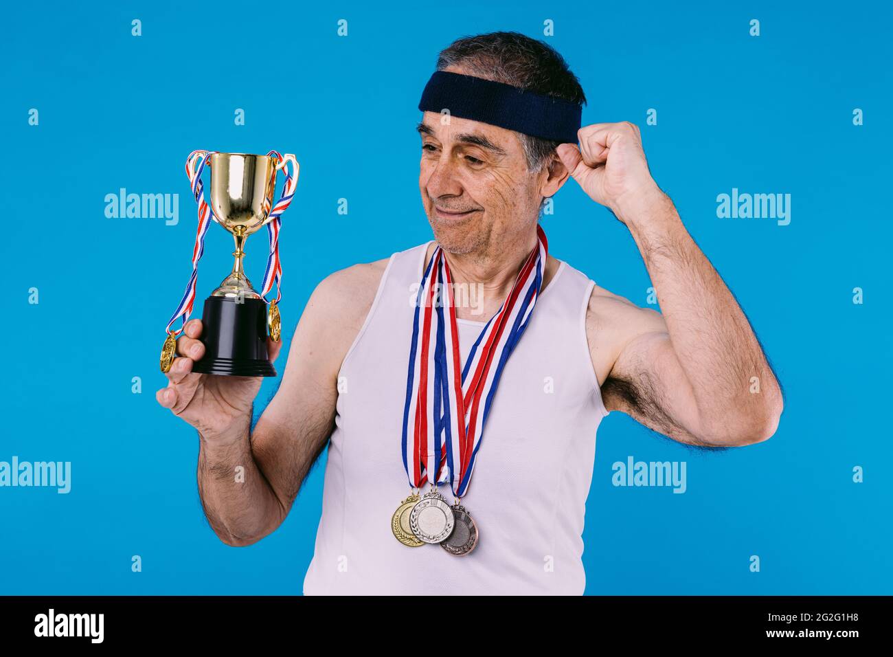 Atleta anziano con marcature solari sulle braccia, con tre medaglie sul collo e un trofeo nelle mani, aggrappando il pugno come segno di forza, su blu Foto Stock
