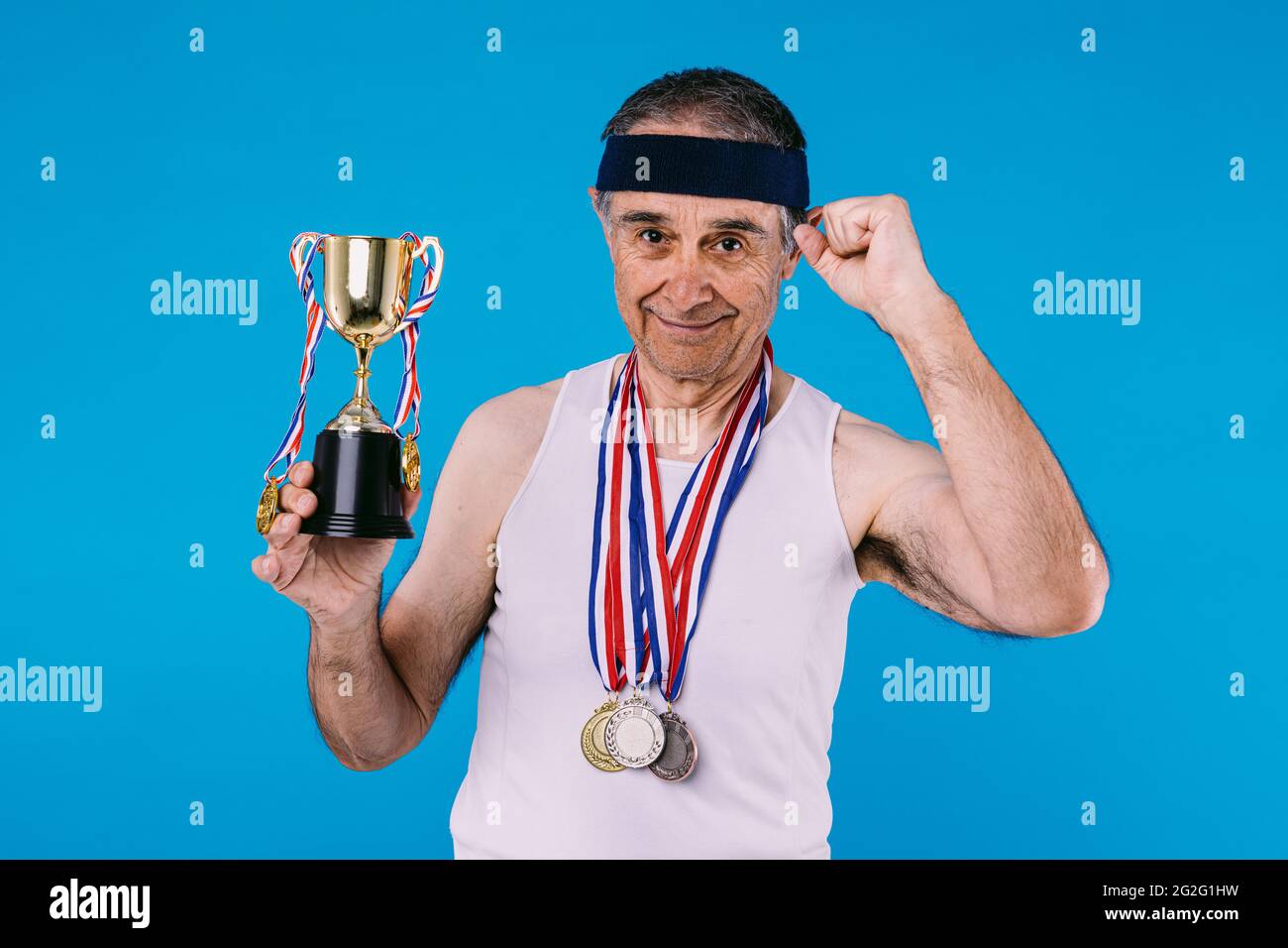 Atleta anziano con marcature solari sulle braccia, con tre medaglie sul collo e un trofeo nelle mani, aggrappando il pugno come segno di forza, su blu Foto Stock