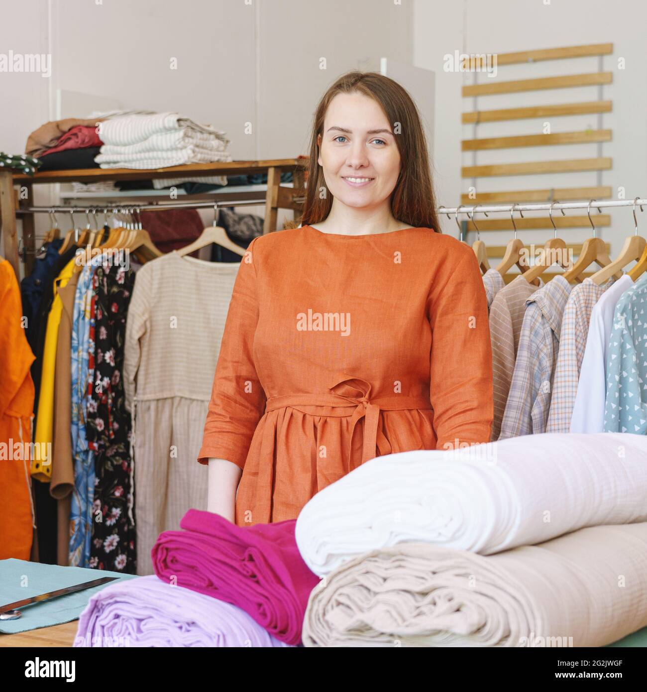 Positivo proprietario di atelier femminile in posa vicino a rotoli di tessuto e abiti in sullo sfondo Foto Stock